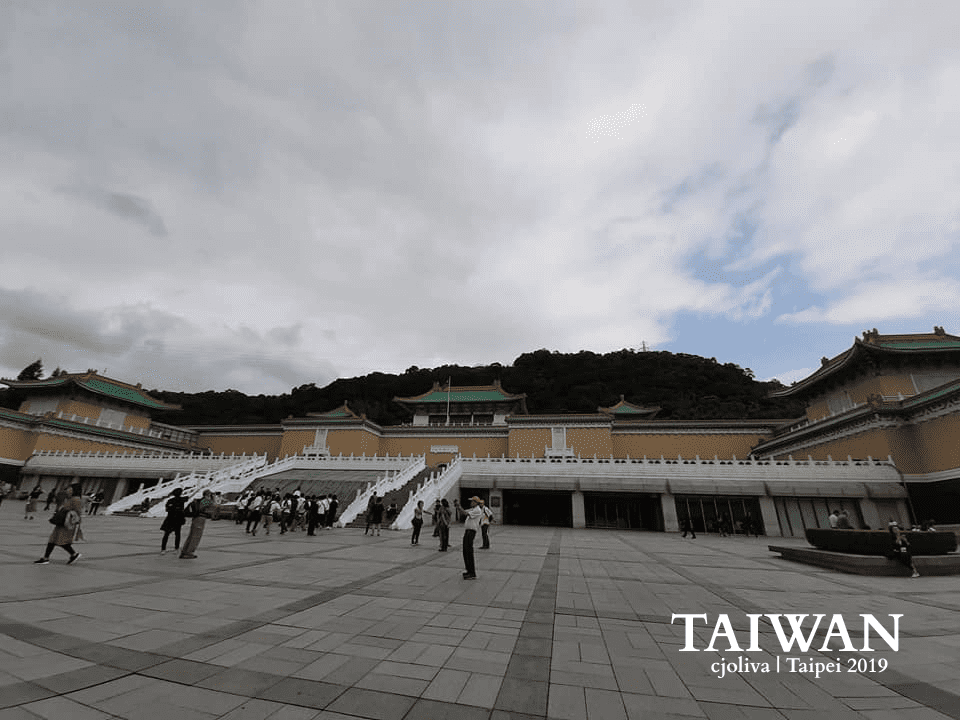 A wide-angle perspective of the National Palace Museum in Taipei, Taiwan. The shot features a grand stone walkway flanked by manicured evergreen hedges, leading toward the iconic palace-style museum building nestled against a lush, forested mountain under a cloudy sky.