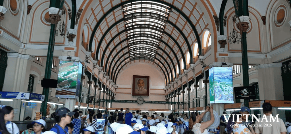 Interior of Saigon Central Post Office in Ho Chi Minh City with arched glass ceiling, ornate cream and orange walls, green columns with gold accents, and a portrait of Ho Chi Minh above a clock.