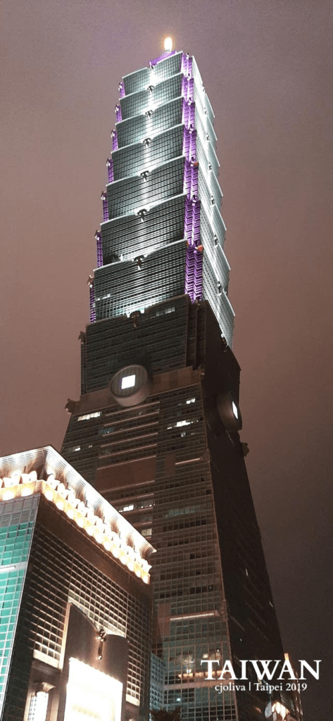 A vertical night shot of the Taipei 101 skyscraper in Taiwan, featuring vibrant purple lights on its upper tiers against a dark sky, highlighting its pagodalike architecture.