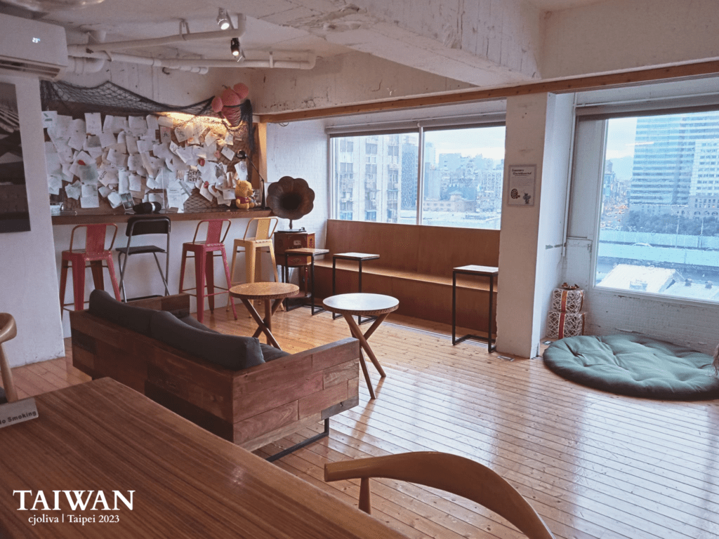 A warm, sunlit interior of a Taipei cafe featuring polished wooden floors, a rustic wooden sofa, and high bar stools. A large window offers a city view, while a wall decorated with notes and a vintage gramophone adds a creative, bohemian touch to the space.