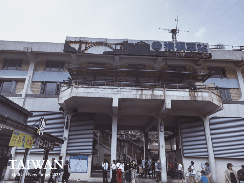 Exterior of Houtong Station in New Taipei, Taiwan with concrete facade, signage in Chinese and English, crowds entering and exiting, colorful shop signage, and rust‑stained textures.
