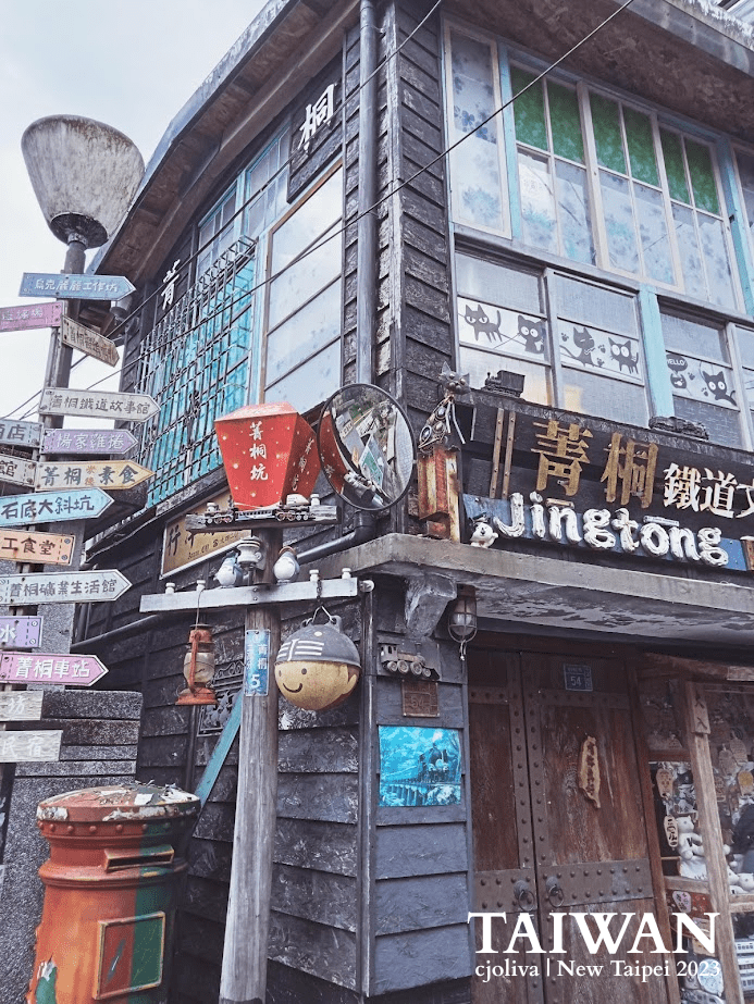 Rustic wooden building in Jingtong, New Taipei, Taiwan, with weathered panels, tinted glass windows, Chinese and English signage reading “Jingtong,” a red lantern, figurines, a vintage mailbox, and colorful directional signs pointing to local destinations.