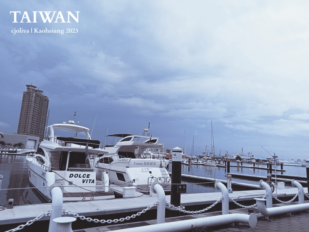 A solo travel experience from Kaohsiung to Taipei Marina in Kaohsiung, Taiwan, with yachts including Dolce Vita and Urania docked along a calm waterfront, a tall building in the background, and masts and boats lining the harbor under an overcast sky.