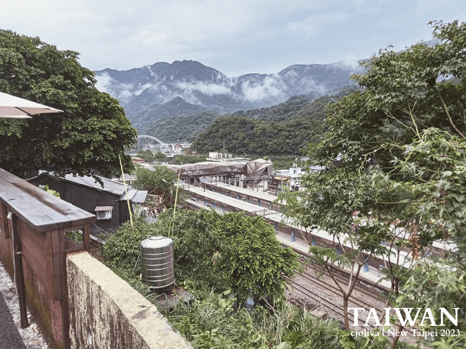 Scenic railway station in New Taipei, Taiwan, surrounded by lush green hills and dense vegetation, with wooden railings, a concrete wall, a metal water tank in the foreground, train tracks, and station platforms in the midground, and misty forested mountains under a cloudy sky.