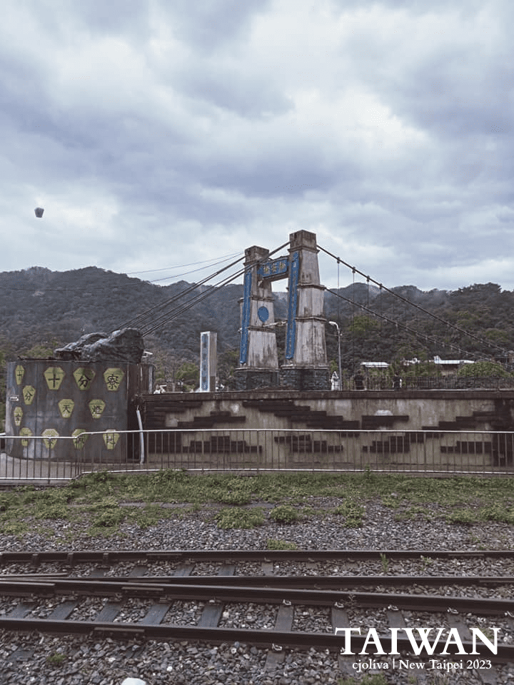 Suspension bridge in New Taipei, Taiwan with weathered concrete towers painted blue, steel cables, railway tracks running parallel, decorative wall with yellow shield symbols, lush green hills, and lantern floating in cloudy sky.