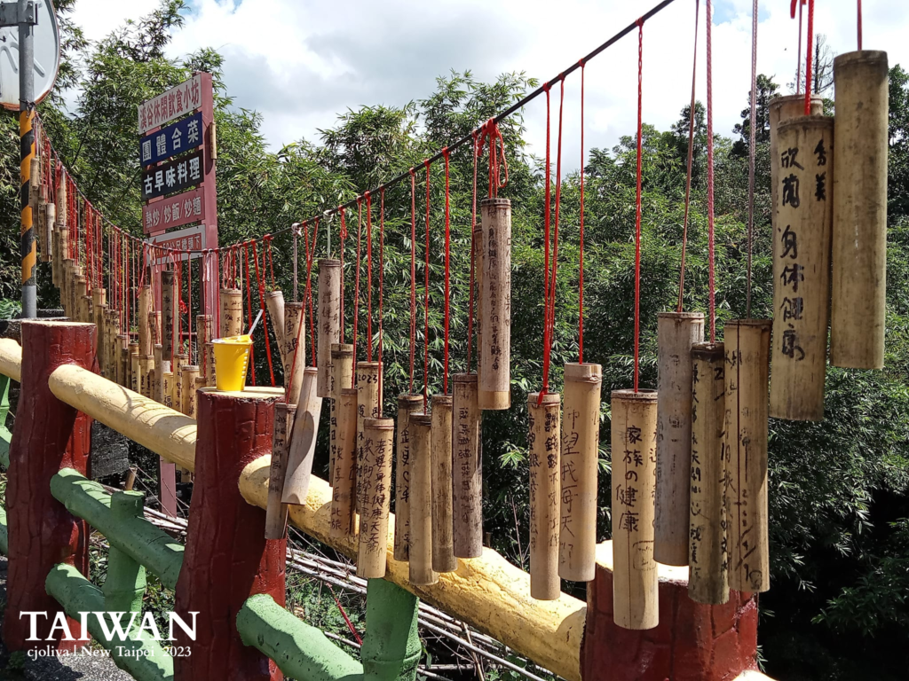Outdoor display in Pingxi, New Taipei, Taiwan, with bamboo tubes hanging from red strings on a colorful railing, each inscribed with handwritten wishes, surrounded by lush greenery and local tea signage.