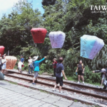 A group of people in Pingxi, New Taipei, Taiwan, releasing colorful sky lanterns—red, orange, white, and blue—each inscribed with handwritten wishes, along railway tracks surrounded by greenery.