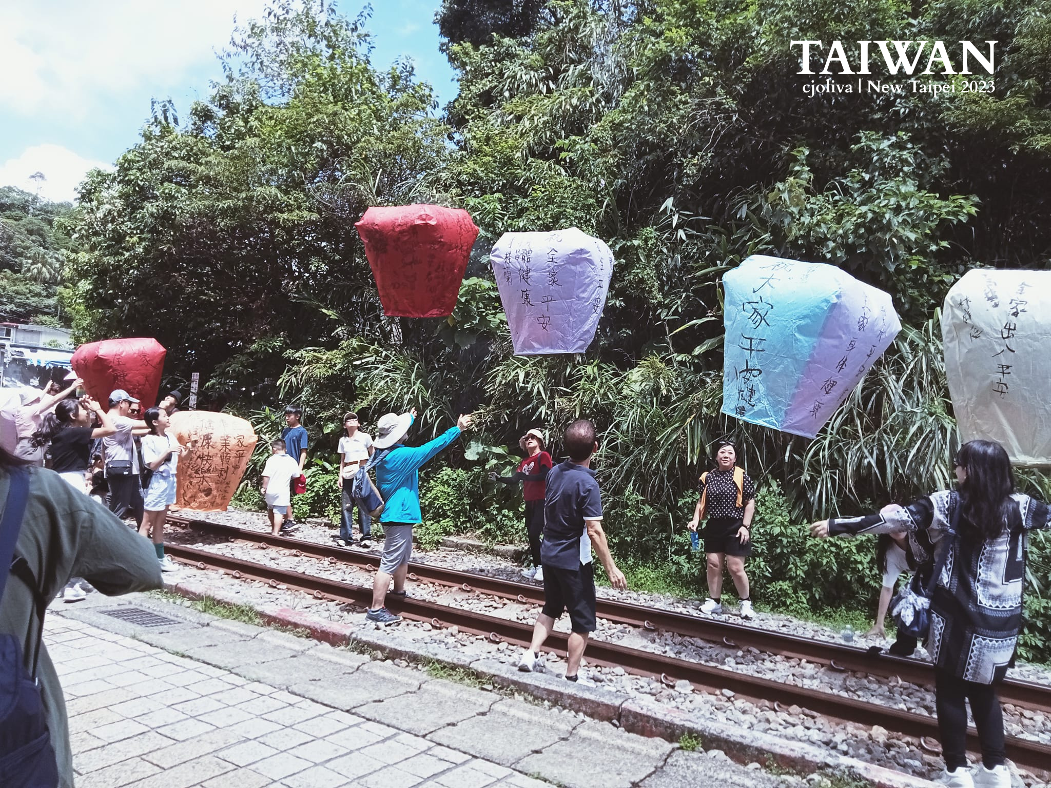 A group of people in Pingxi, New Taipei, Taiwan, releasing colorful sky lanterns—red, orange, white, and blue—each inscribed with handwritten wishes, along railway tracks surrounded by greenery.