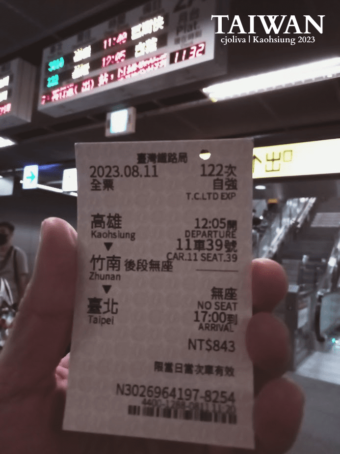 A hand holding a Taiwan Railways (TRA) T.C. Ltd. Exp. train ticket dated August 11, 2023. The ticket shows a journey from Kaohsiung to Taipei via Zhunan, with a departure time of 12:05 and a price of NT$843, set against a blurred station departure board.
