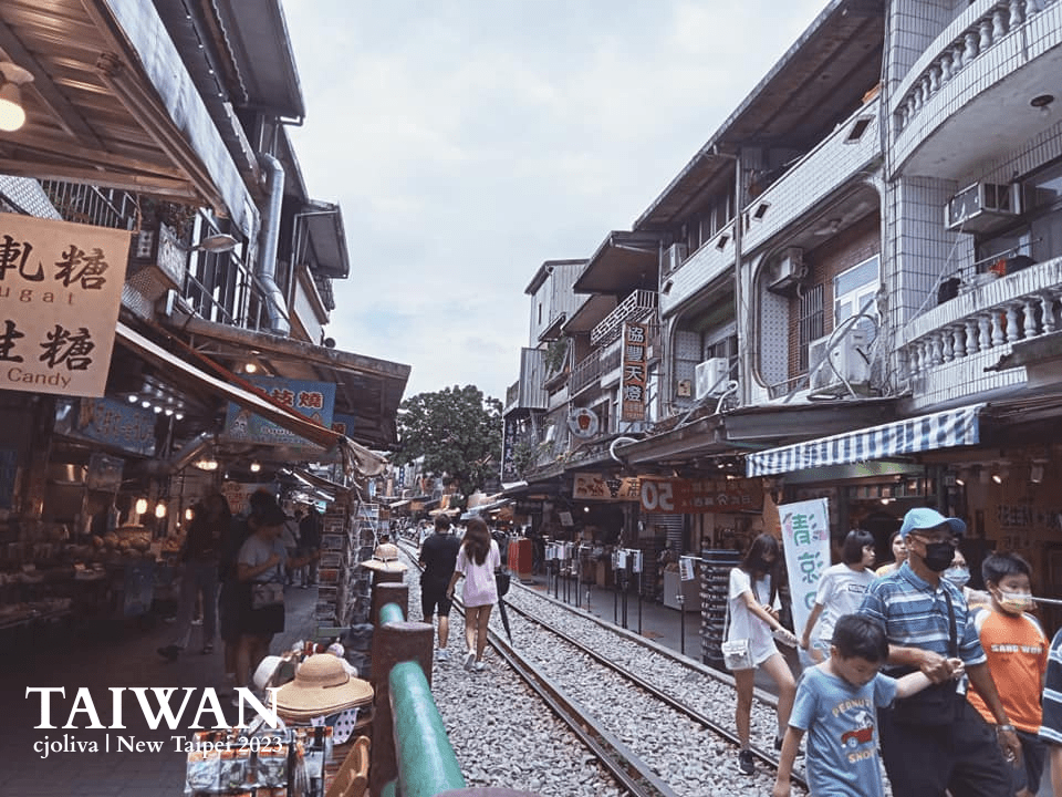 Street scene at Shifen Old Street in New Taipei, Taiwan, with railway tracks running through the center, traditional buildings with tiled exteriors and balconies, shops and stalls, and people walking along the tracks.