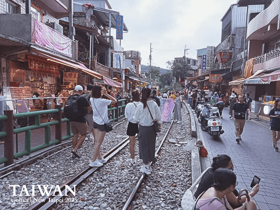 Street scene at Shifen Old Street in New Taipei, Taiwan with railway tracks running through a busy market, people walking and taking photos, colorful sky lanterns, shop signs, and scooters parked along the street.