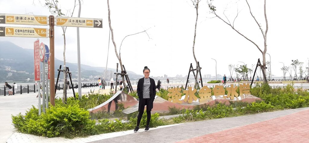 A woman standing in front of the "Tamsui Golden Anchor" sign at the waterfront park in New Taipei City, Taiwan, with directional signs for the MRT and Ferry Wharf in the background.