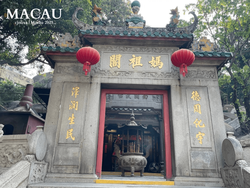 A view of the stone entrance gate to the A-Ma Temple in Macau, decorated with red lanterns, gold Chinese inscriptions, and a large bronze incense burner in the center.