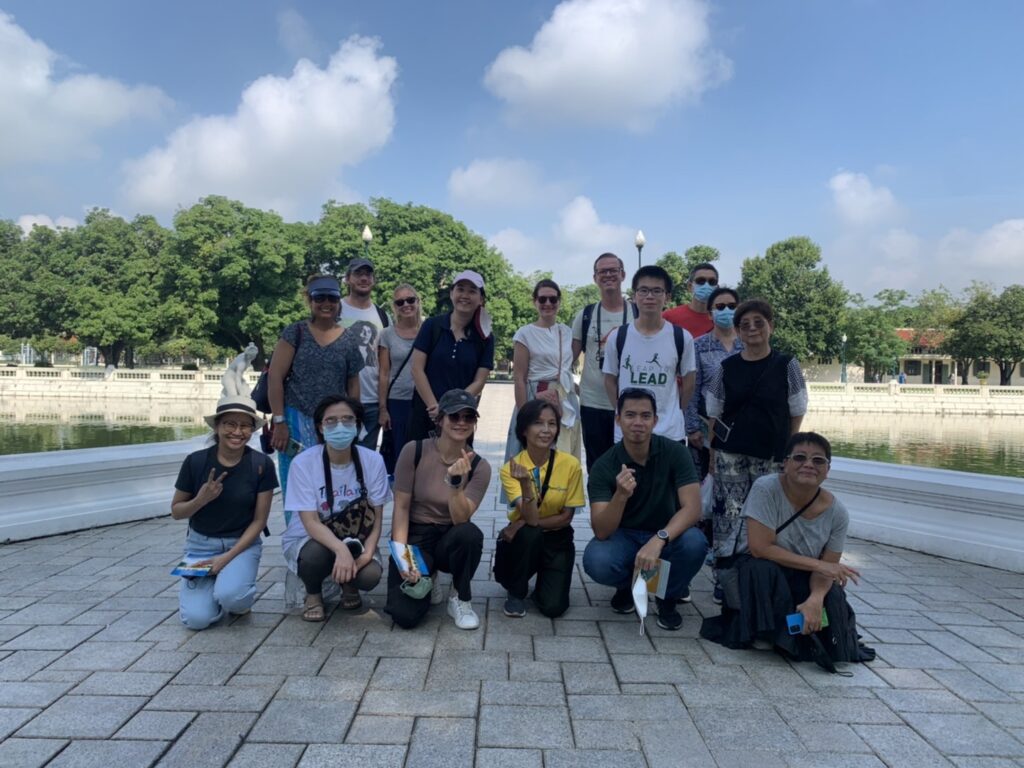 A group of 17 people posing near a body of water with trees, railing, and statue in Ayutthaya, Thailand