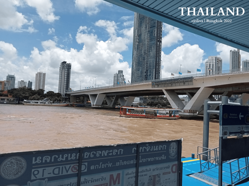 Passenger boat traveling under a bridge with Thai flags along the Chao Phraya River in Bangkok, surrounded by high-rise buildings