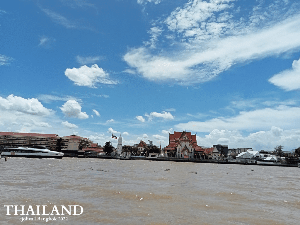 Traditional Thai temple with red roof along the Chao Phraya River in Bangkok, large boat passing by under a bright sky with Thai flag visible