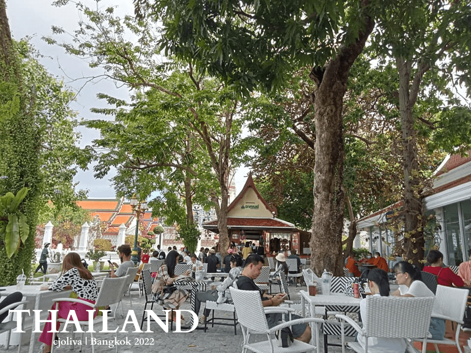 Outdoor café in Bangkok shaded by trees with wicker tables and chairs, people dining near temple rooftops in the background