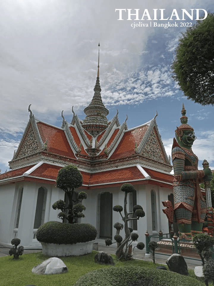 Ornate Thai temple with multi-tiered red and gold roof, trimmed topiary trees, and colorful guardian statue under a partly cloudy sky