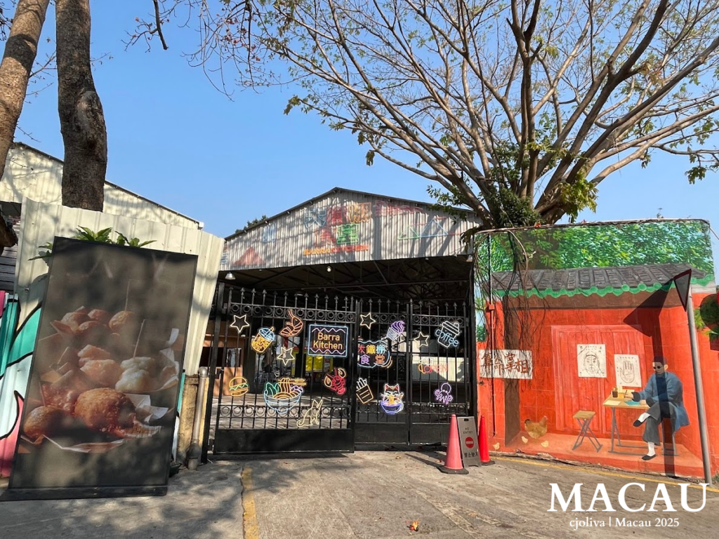 The entrance to an industrial-style food and art hub in Macau, featuring black iron gates decorated with colorful neon icons of food and stars, flanked by a large mural of a traditional scholar on a red background.