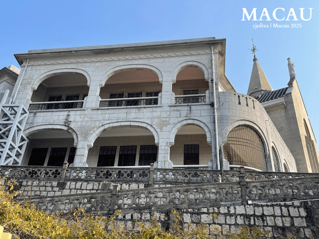 A side view of the grey stone Chapel of Our Lady of Penha in Macau, showing its arched balconies, stone railings, and the pointed spire of the bell tower against a blue sky.