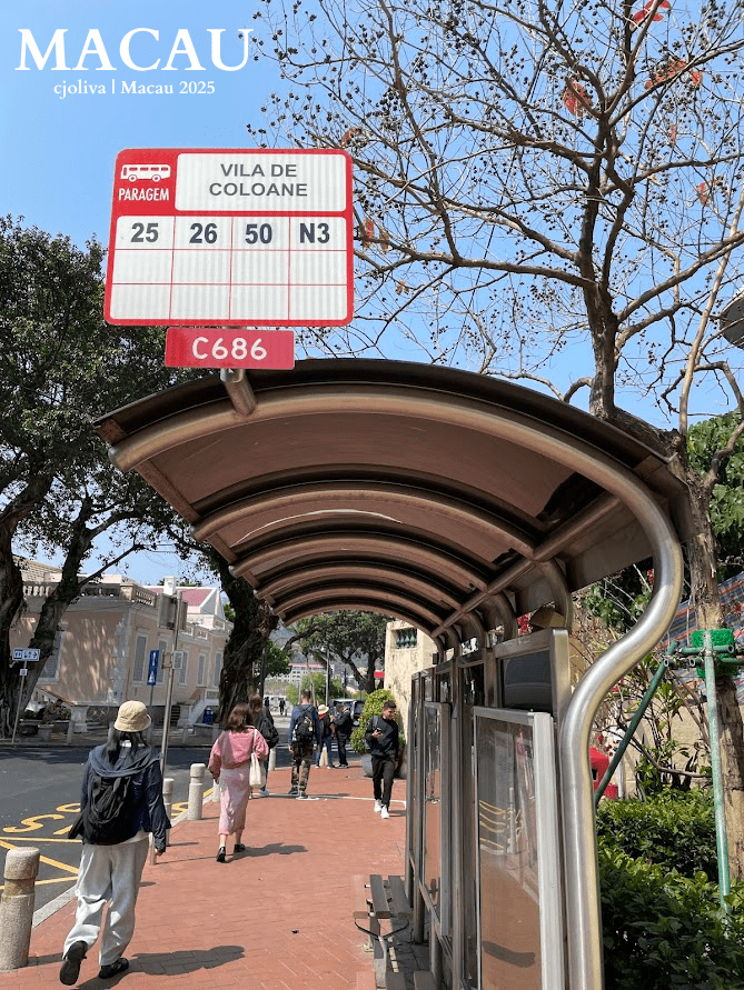 A red and white bus stop sign for "Vila de Coloane" showing routes 25, 26, 50, and N3, standing over a modern arched waiting shelter on a brick sidewalk with people walking by.