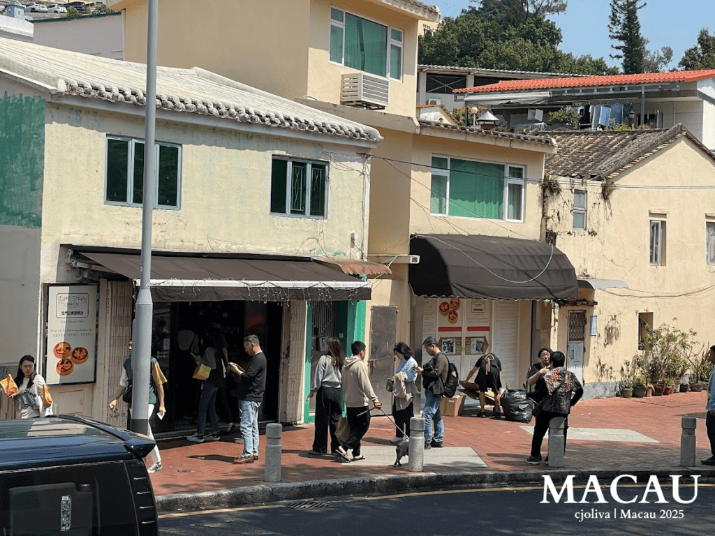 A street-side view of the rustic, small storefront of Lord Stow’s Bakery in Coloane Village, featuring its iconic yellow signage and people gathered outside to buy fresh egg tarts.