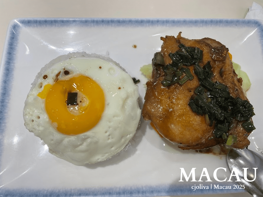 Plated birthday dinner at Macau International Airport featuring rice topped with a sunny-side-up egg, grilled chicken with sauce and green onions, and fresh vegetables.