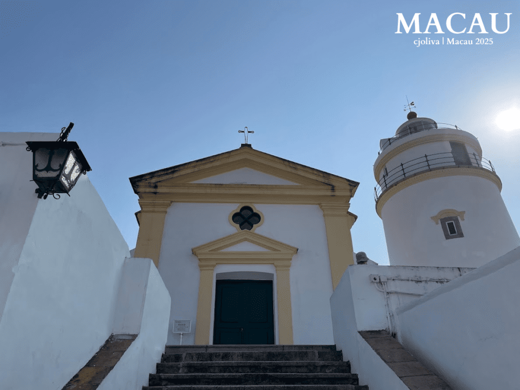 Low-angle shot looking up at the white and yellow Guia Chapel and the adjacent circular Guia Lighthouse under a bright sun at the top of a stone staircase.