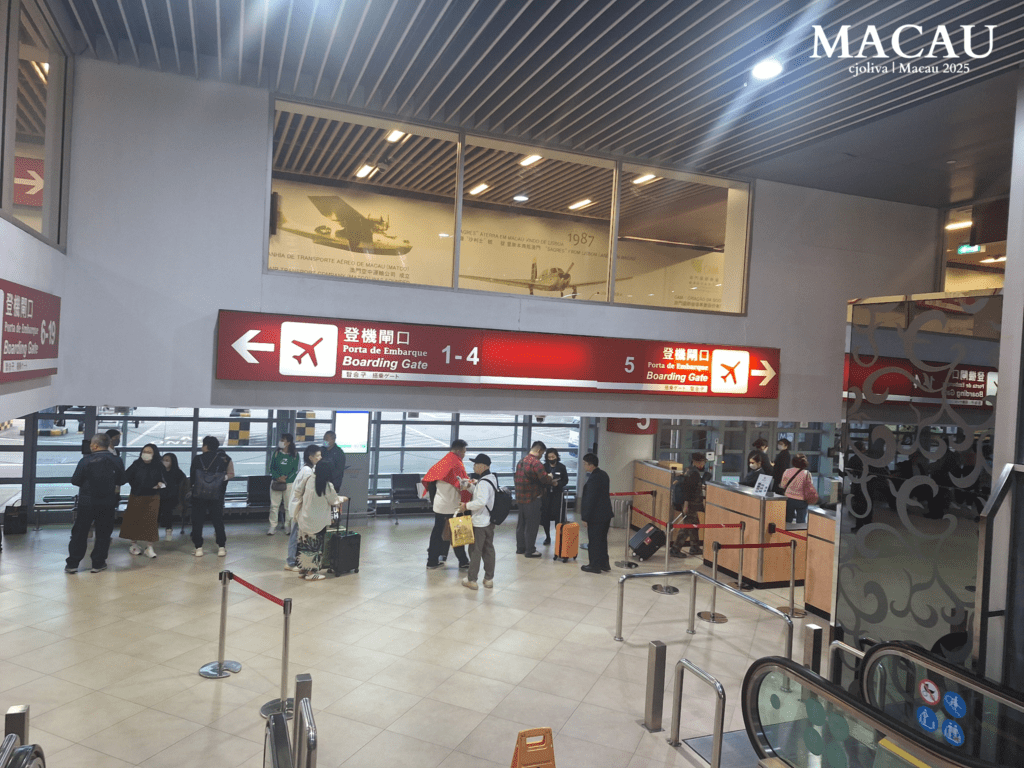 Passengers at Macau International Airport boarding area with multilingual gate signs, modern counters, and a historical display referencing the airport’s 1987 inauguration.
