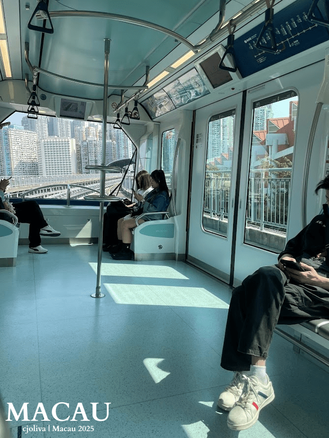 The clean, modern interior of a Macau LRT train car with bright blue and white seating, stainless steel grab bars, and large panoramic windows showing the Barra station platform.