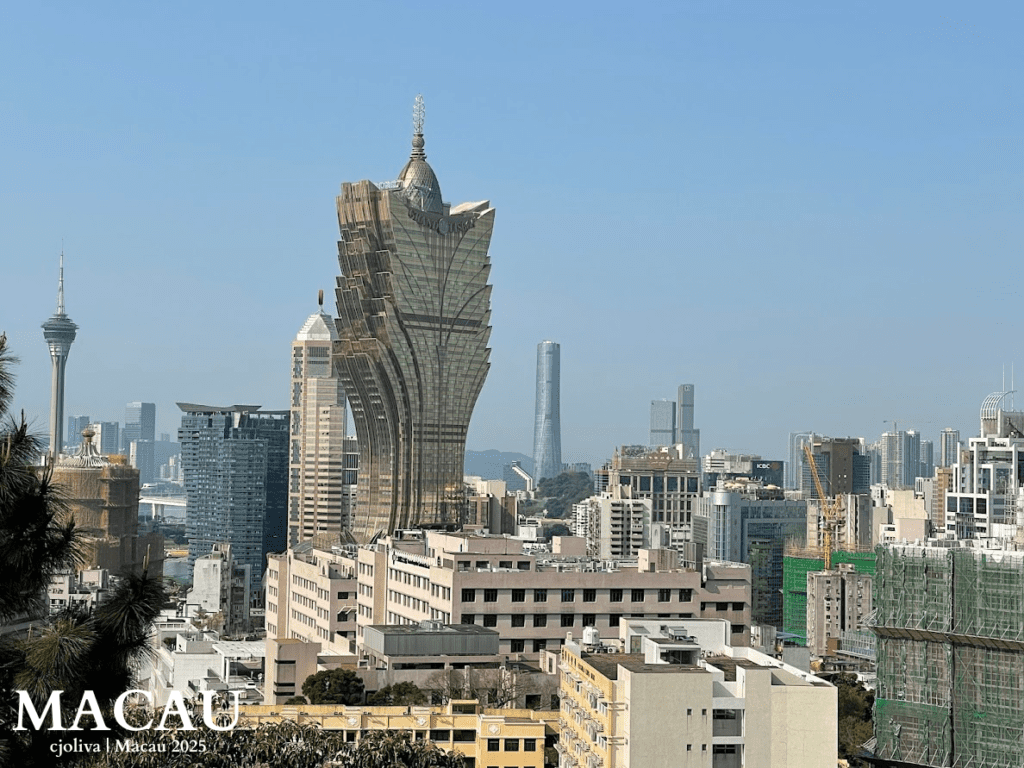 A wide cityscape of Macau featuring the lotus-shaped Grand Lisboa hotel and the slender Macau Tower under a clear sky, with various urban buildings and construction in the foreground.