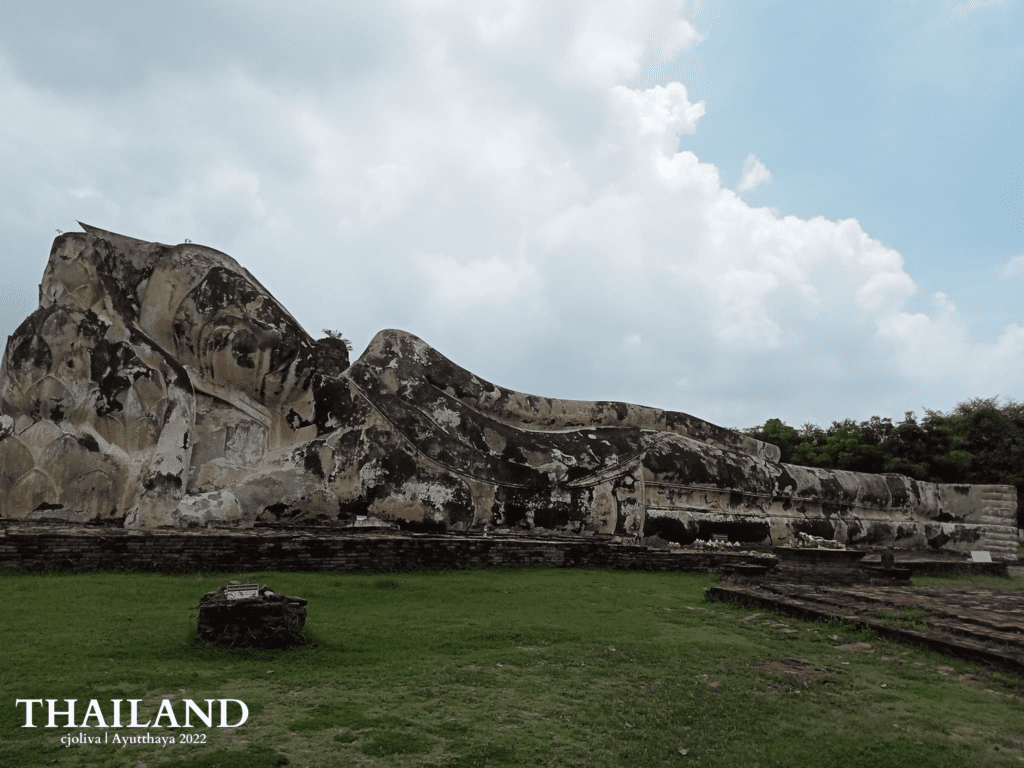 A massive, ancient stone statue of a reclining Buddha resting on its side against a backdrop of white clouds and green trees at the Wat Lokaya Sutha ruins in Ayutthaya, Thailand.