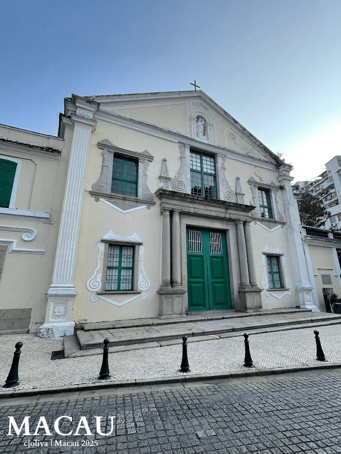 A low-angle shot of the cream-colored neoclassical facade of St. Augustine’s Church in Macau, featuring green doors and windows, white decorative pillars, and a cobblestone street in the foreground.