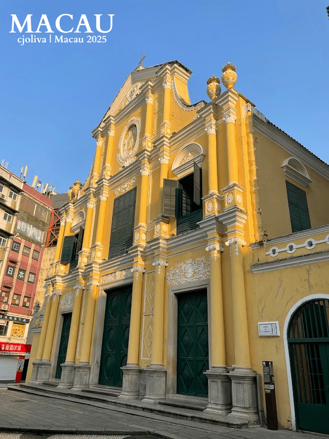 A low-angle shot of the bright yellow Baroque facade of St. Dominic's Church in Macau, featuring white stucco ornaments and green shutters under a clear sky.