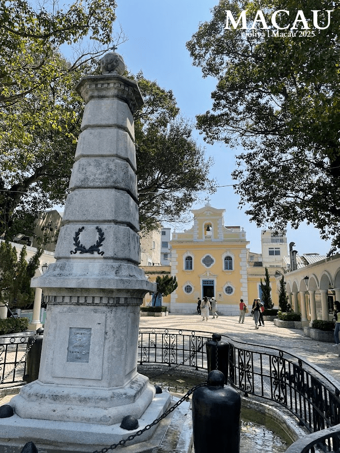 A white stone monument with a laurel wreath stands in a sunny square paved with traditional Portuguese wave-patterned stones. In the background is the bright yellow St. Francis Xavier Chapel under a clear sky.