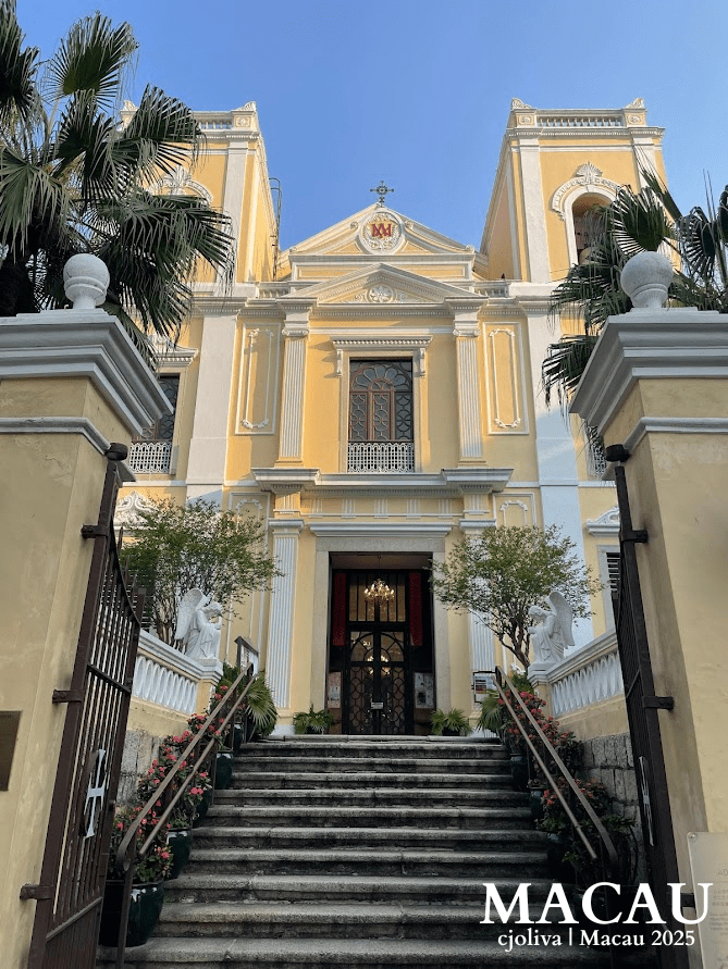 A symmetrical shot looking up the stone steps toward the yellow and white neoclassical facade of St. Lawrence's Church, flanked by palm trees and angel statues.