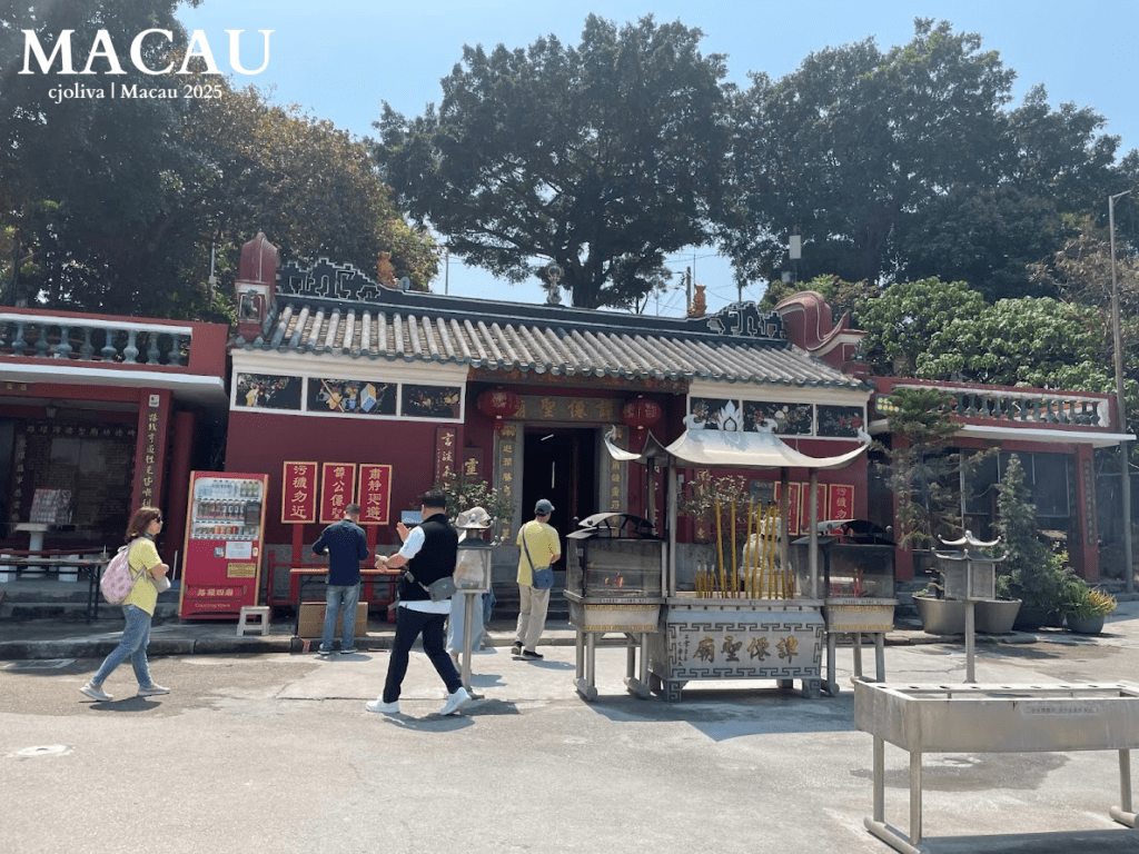 A wide shot of the red-walled Tam Kung Temple with traditional grey-tiled roofing and colorful ceramic figures. A large incense burner stands in the courtyard with visitors paying their respects.