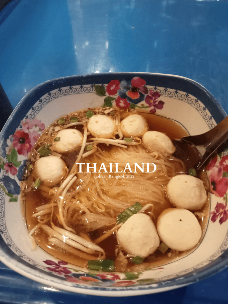 Bowl of Thai noodle soup with rice noodles, fish balls, bean sprouts, and green onions in dark broth on a blue table