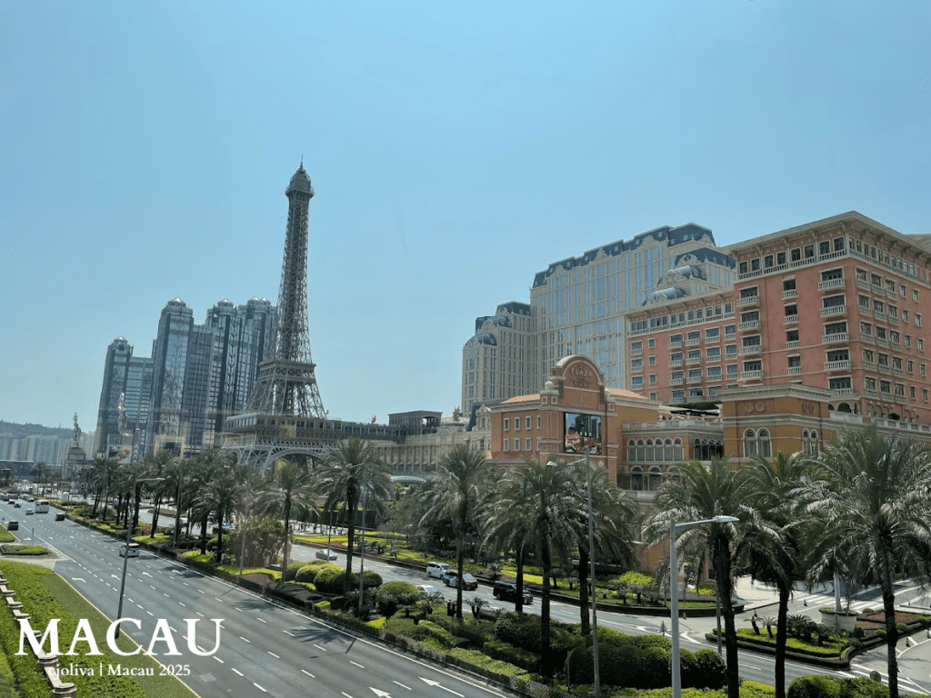 A wide landscape view of the half-scale Eiffel Tower replica at The Parisian Macao, surrounded by palm trees and luxury hotel buildings under a bright sky.