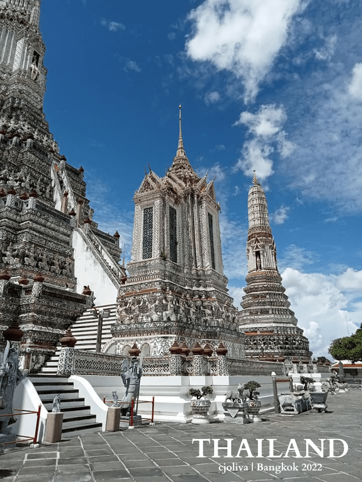 Intricately decorated prangs at Wat Arun temple in Bangkok with colorful porcelain details, stone statues, and staircases under a bright blue sky