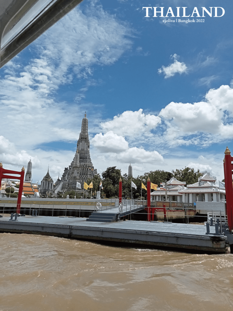 Wat Arun temple with central prang rising above the Chao Phraya River, dock and flags in the foreground under a bright sky