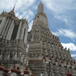 Ornate Wat Arun temple in Bangkok with towering prang decorated in porcelain under a partly cloudy sky