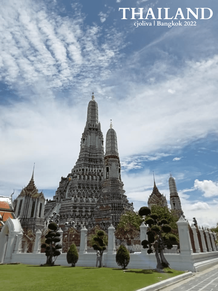 Wat Arun temple in Bangkok with central prang, manicured lawn, sculpted trees, and ornate Thai architecture under a partly cloudy sky