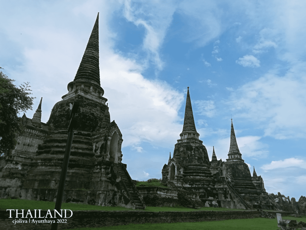 Three large, weathered, bell-shaped stone chedis stand in a row against a blue sky with soft white clouds, surrounded by green grass and the brick foundations of the ancient Royal Palace in Ayutthaya.