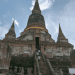 A view from the ground up of the massive, central, bell-shaped chedi at Wat Yai Chai Mongkhon in Ayutthaya, Thailand, flanked by two smaller chedis under a dramatic, cloudy sky.