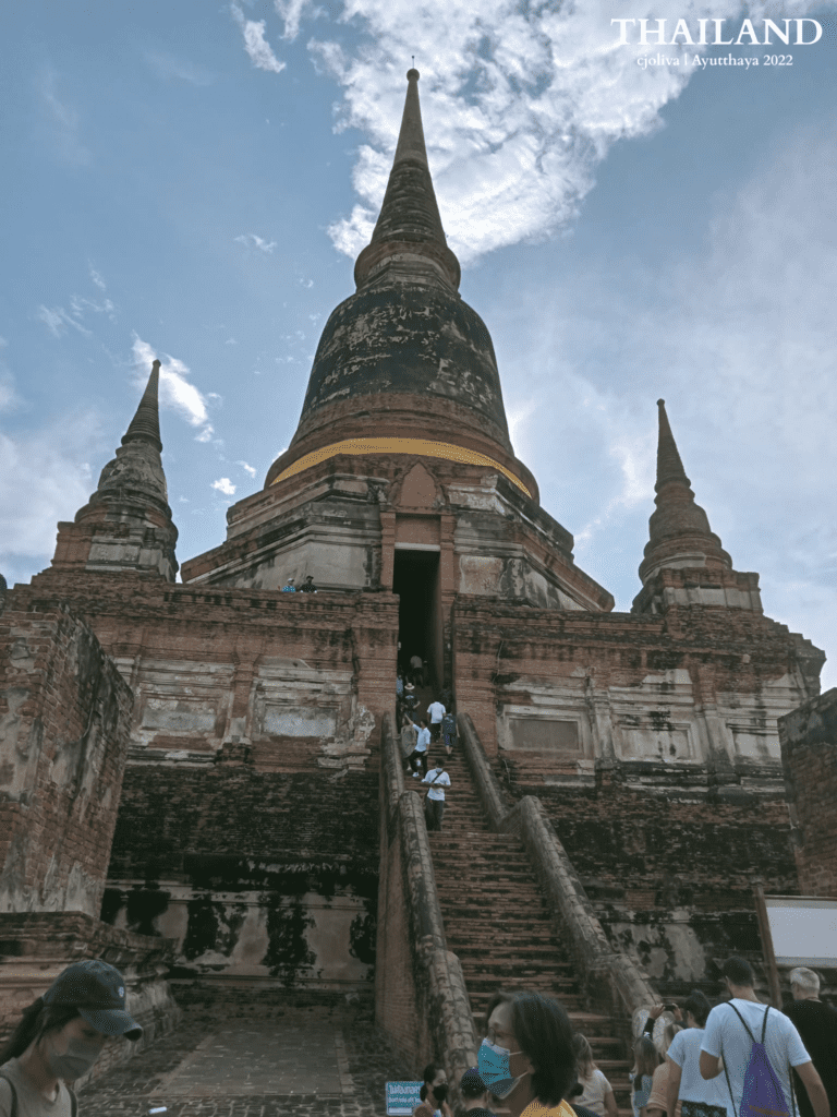 A view from the ground up of the massive, central, bell-shaped chedi at Wat Yai Chai Mongkhon in Ayutthaya, Thailand, flanked by two smaller chedis under a dramatic, cloudy sky.