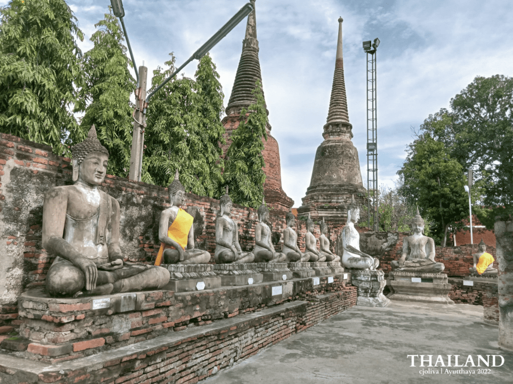 A row of serene, weathered stone Buddha statues in a meditative seated position, some draped in traditional yellow sashes, lined up against an ancient red-brick wall with towering chedis in the background.