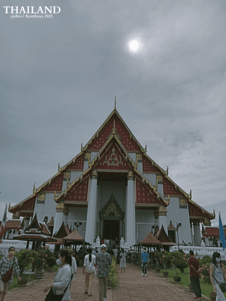 A symmetrical shot of the white and red Wihan Phra Mongkhon Bophit temple under a cloudy sky with the sun visible. People walk along the brick path leading to the entrance of the grand assembly hall in Ayutthaya.