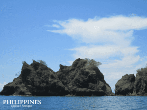 Scenic coastal view in Antique, Philippines, with rugged rock formations rising from the sea, sparse vegetation, calm blue water, and bright sky with scattered clouds.