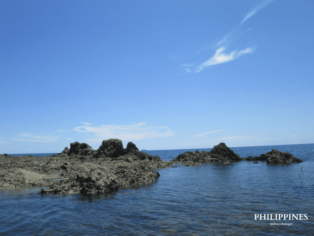 Scenic coastal view in Antique, Philippines, with rugged dark rock formations extending into calm blue sea under a clear sky with scattered clouds, taken in 2018
